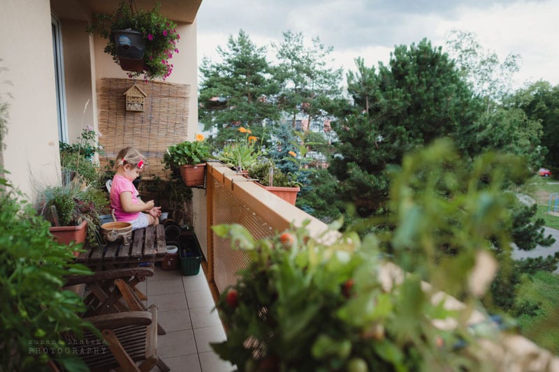 Balkon plný kytek, uprostřed malá postava v růžové